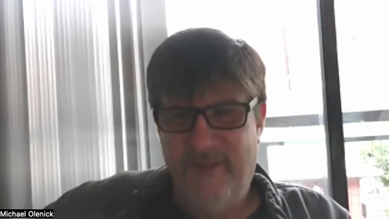 Well-centered, clear webcam screenshot of a presenter speaking in front of a bright window and vertical blinds.