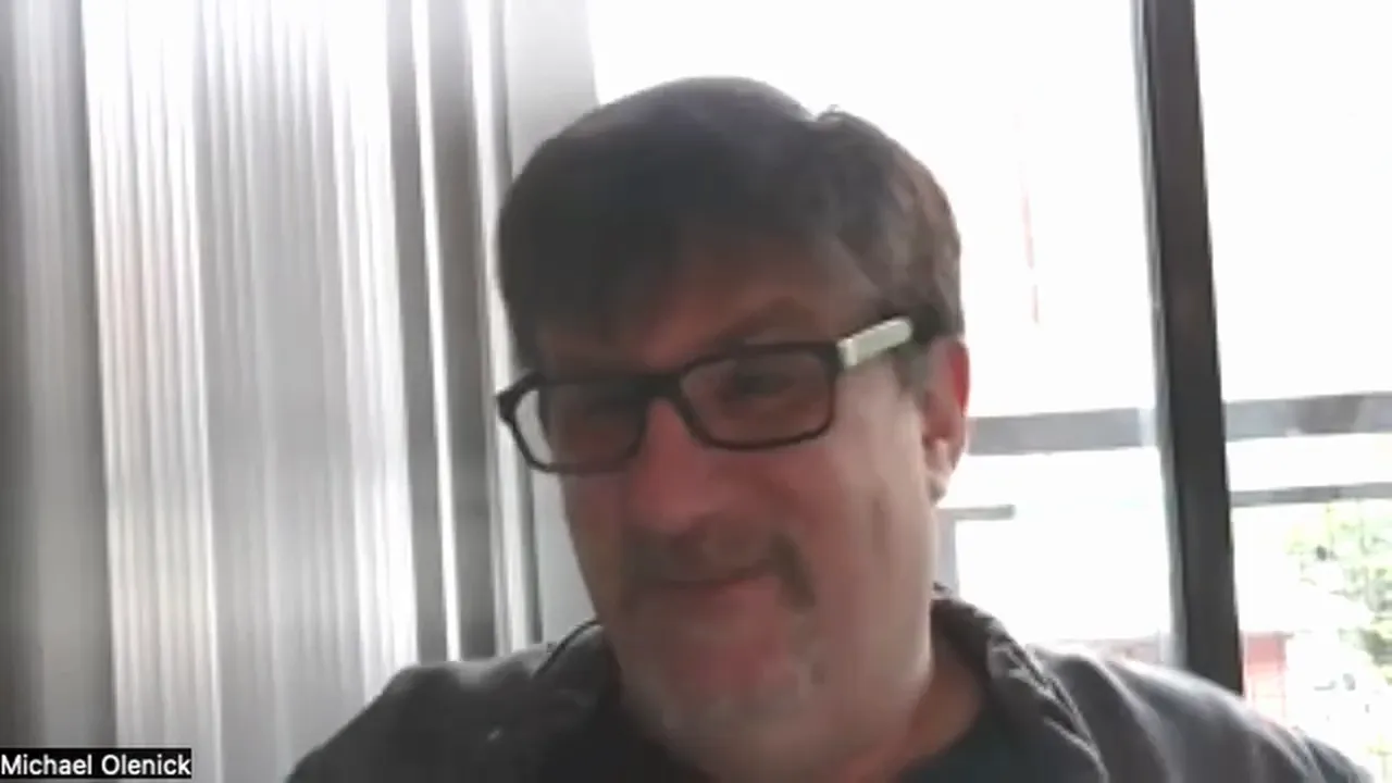 well-framed screenshot of a video-call participant speaking with vertical blinds and a window behind