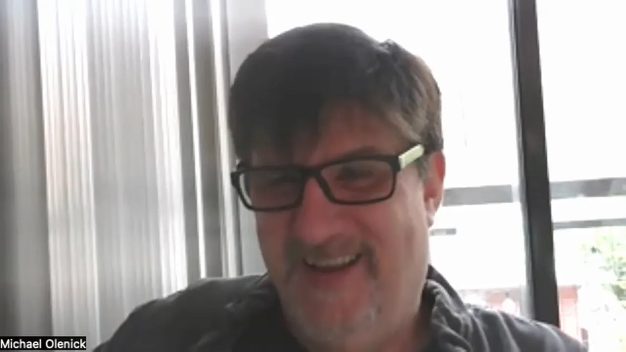 Well‑lit webcam screenshot of a presenter smiling, centered with a bright window and vertical blinds behind.