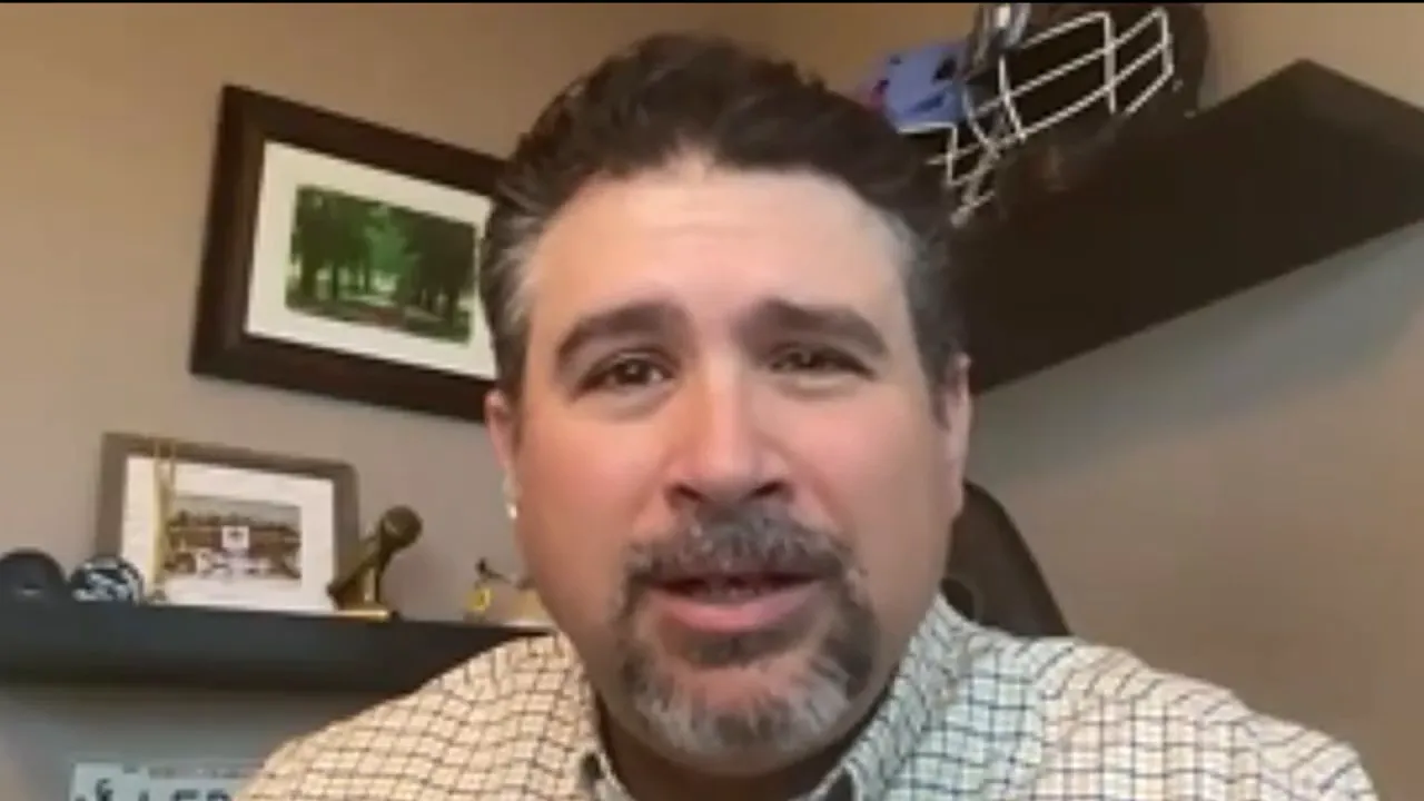 clear videocall screenshot of a man speaking directly to camera with shelves and framed pictures behind him