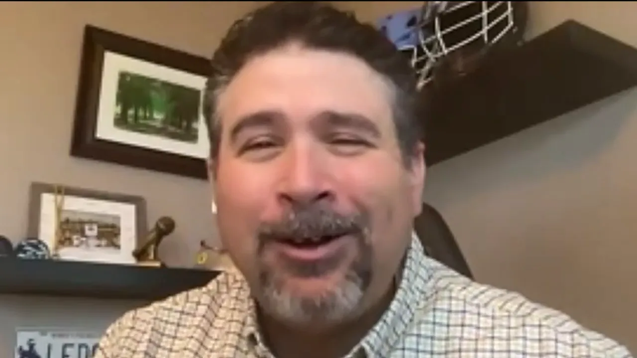 Clear videocall screenshot of a presenter smiling and engaging the camera with home-office shelving behind.