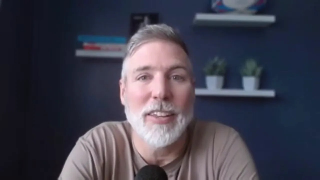 Centered head-and-shoulders portrait of a presenter speaking into a microphone in a home office with navy wall and decorative shelves behind.