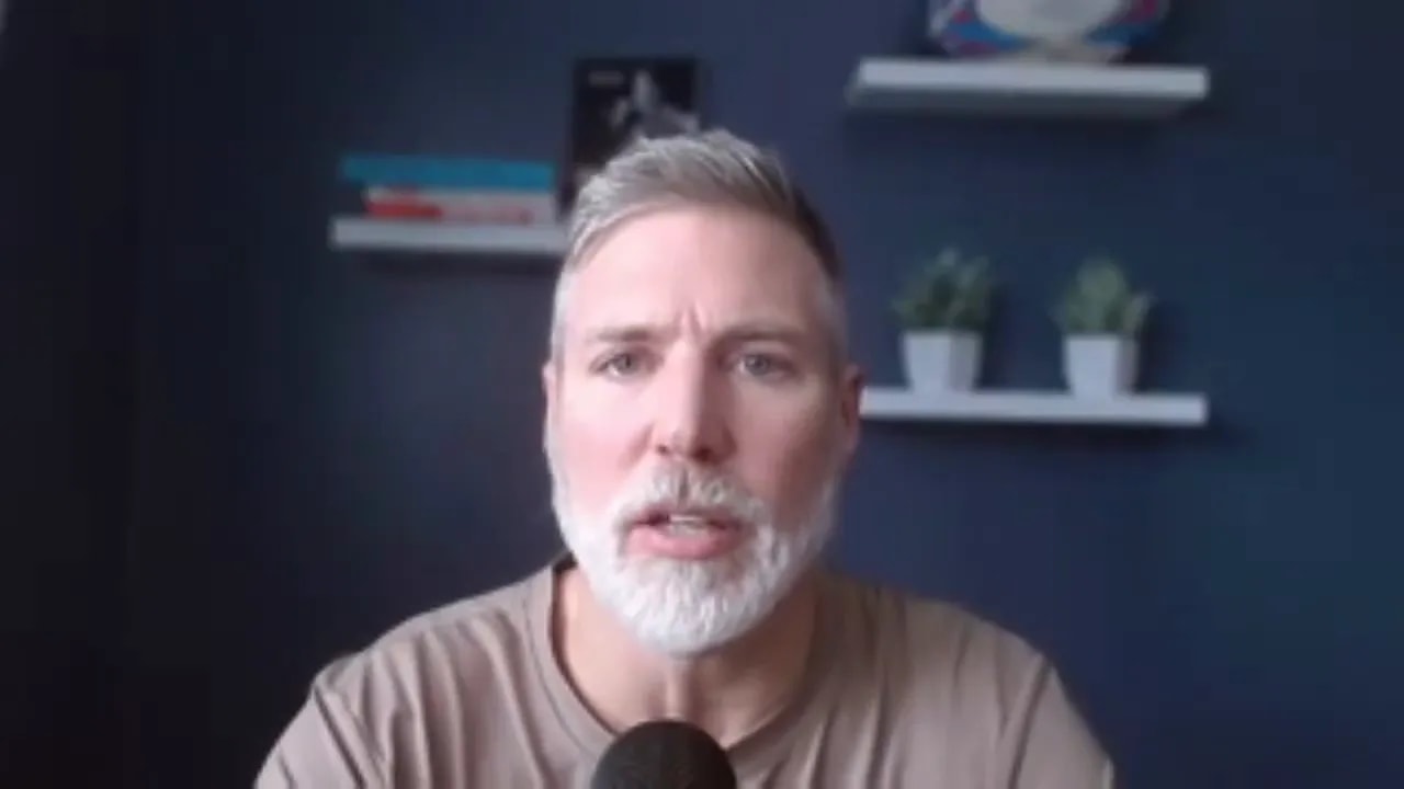 Centered mid-shot of a presenter speaking into a microphone with a navy wall and decorative shelves behind him.