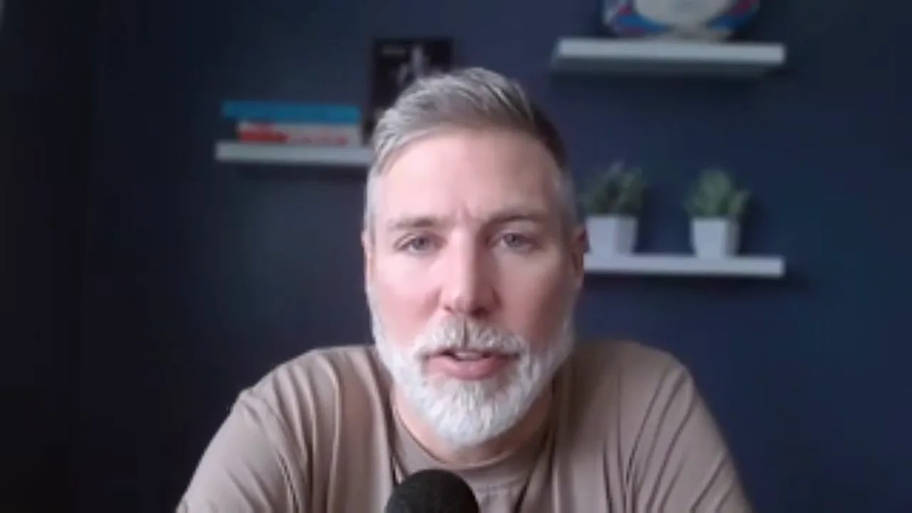 Centered head-and-shoulders shot of a speaker addressing the camera from a home office, microphone visible and shelves on a navy wall behind.