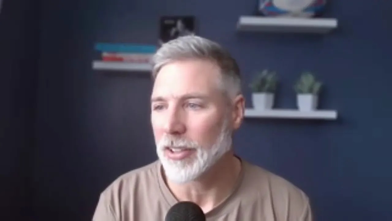 man speaking from a home office with shelves and plants visible behind him