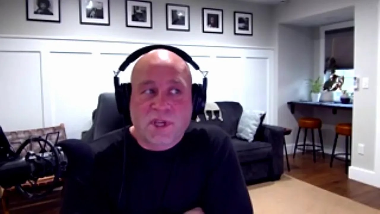 Person wearing headphones speaking into a microphone in a home office, clear view of couch, pillows and counter with stools behind them.