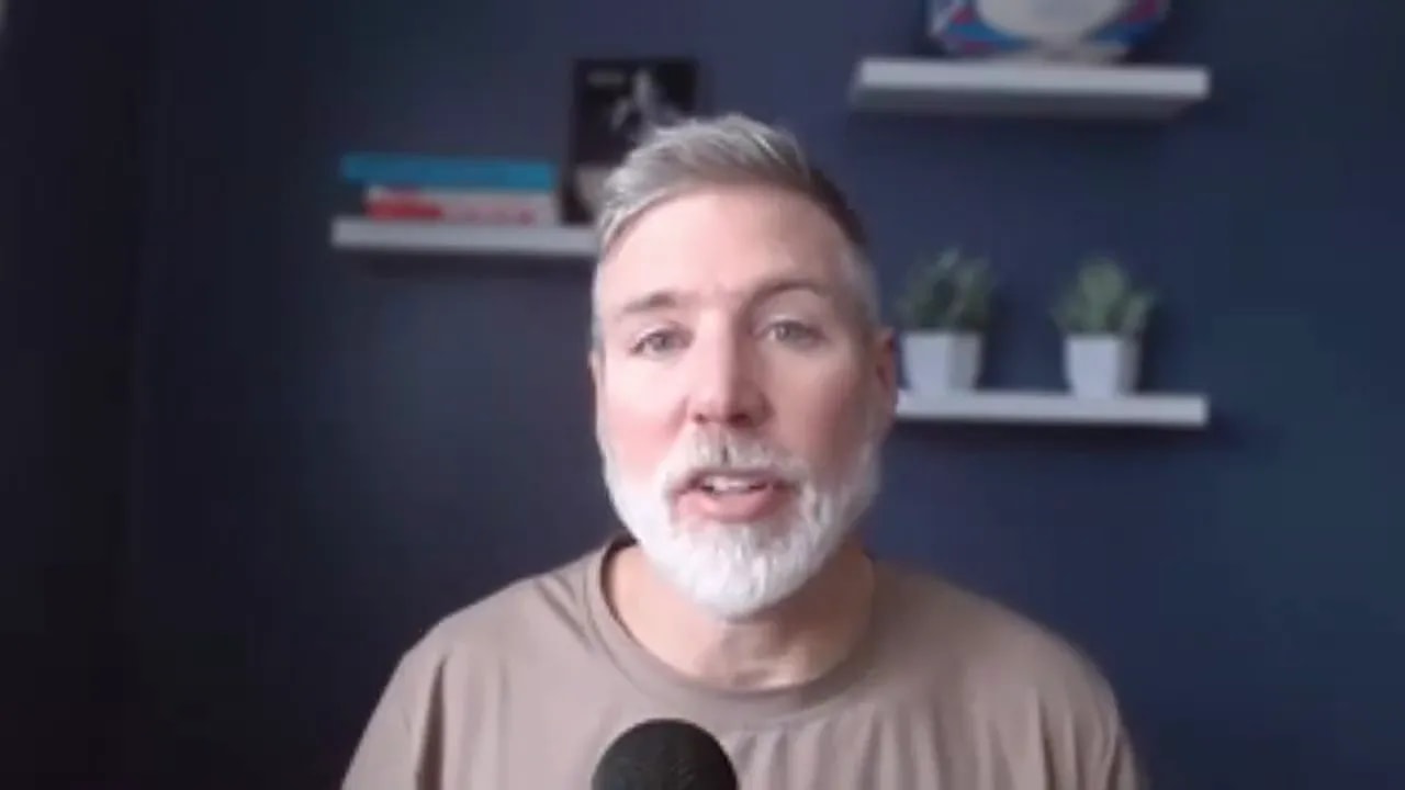 Frontal view of speaker with gray beard speaking into a microphone, clear focus and navy background shelves