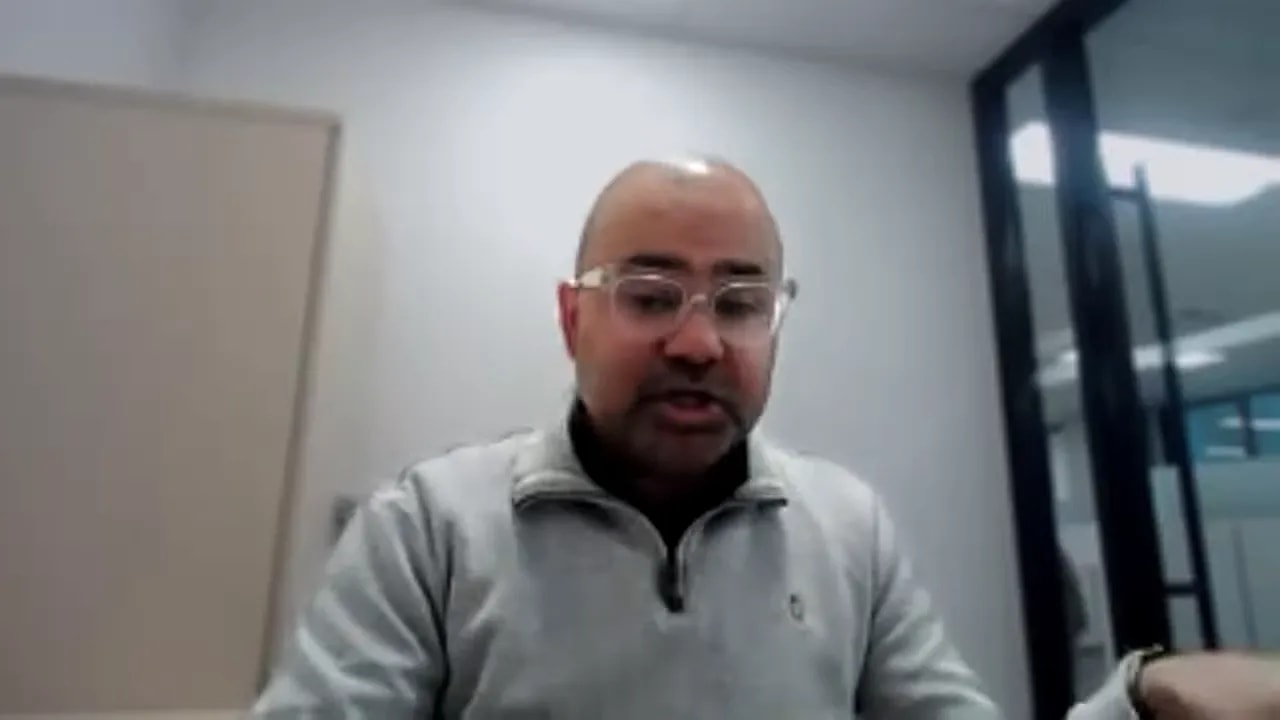 Presenter on a video call in an office, speaking and gesturing with an open hand to the side
