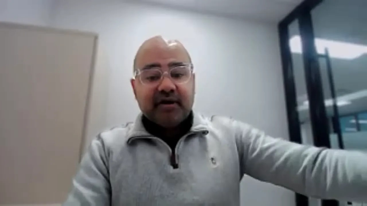 Presenter on a video call in an office, gesturing with an extended arm while explaining funding stacking