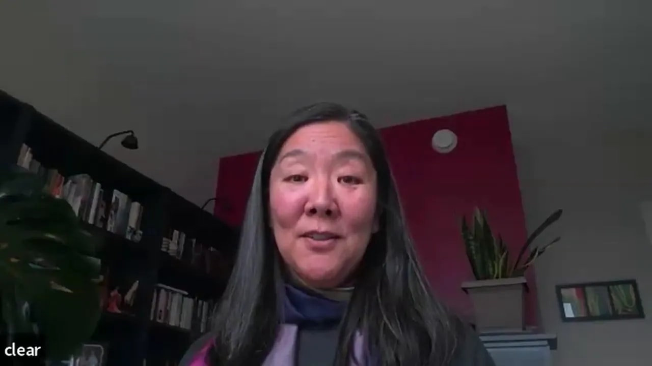 well-lit mid-shot of a leader speaking on a video call with bookshelves and plants visible